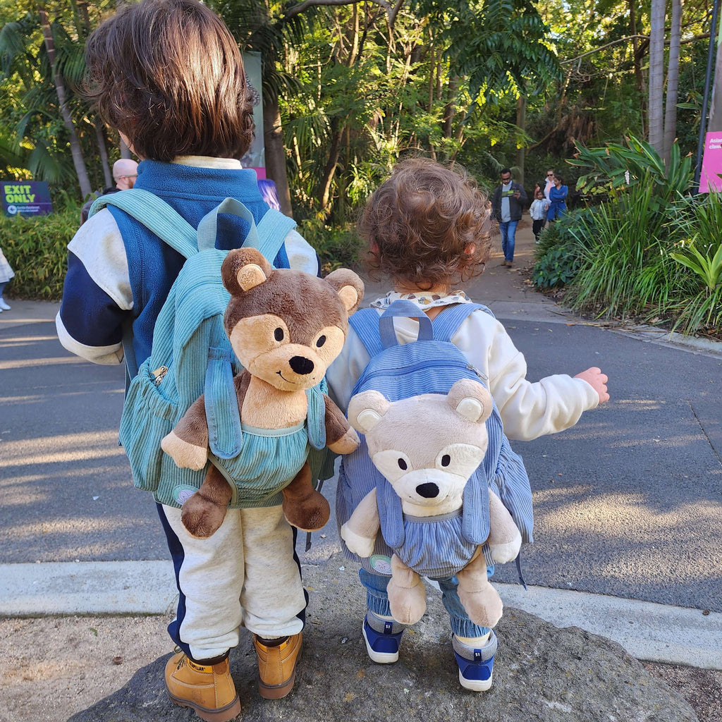 Two children with cudle & carry backpacks featuring teddy bear plush toys, standing on a path in a park-like setting.